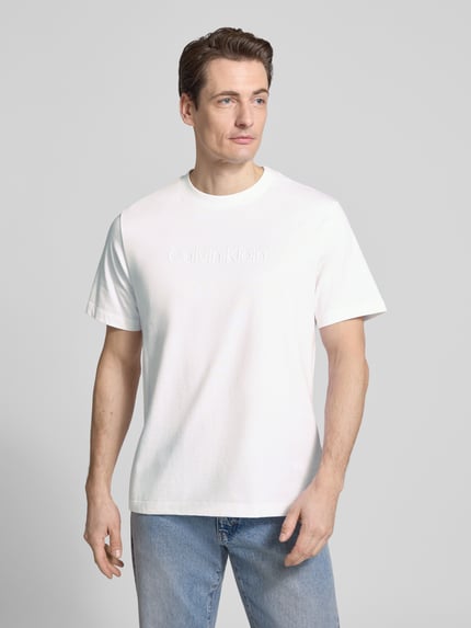 Männermodel trägt T-Shirt mit Rundhalsausschnitt von CK Calvin Klein in Weiß, Herren T-Shirt aus reiner Baumwolle, Regular-Fit, Logo, Ganzkörperansicht von vorne. Rückenlänge bei Größe M: 67 cm.