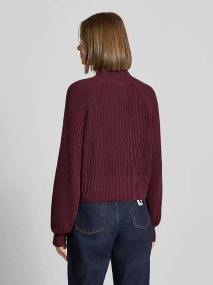 Vrouwenmodel draagt Regular Fit Pullover Baumwoll-Mix Modell 'Dara' van NOISY MAY®, bordeaux, geribde breistructuur, opstaande kraag, strakke zoom, ruglengte 47 cm, mouwlengte 48 cm, strick-look, achteraanzicht.