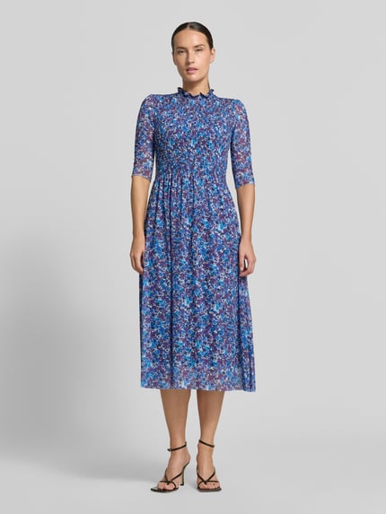 Frauenmodel trägt Midikleid mit Smok-Detail von Rich & Royal, floraler Allover-Print in Blau- und Lilatönen, taillierte Passform, Ganzkörperansicht von vorne. Elastischer Material-Mix, Stehkragen mit Rüschen, halblange Ärmel, Rückenlänge 120 cm, Brustweite 34 cm, Ärmellänge 36 cm.