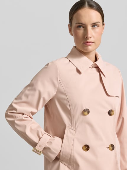 Frauenmodel trägt Trenchcoat mit Knopfverschluss von MICHAEL Michael Kors in zartem Rosa, glatte Textur, Nahaufnahme Oberkörper. Regular Fit mit Umlegekragen, Zweireiher, Details zu Rückenlänge, Schulter- und Brustweite sowie Ärmellänge laut Kontext.