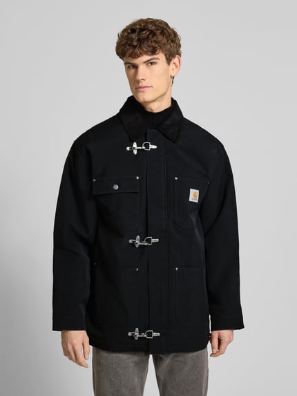 Mannenmodel draagt Oversized Jacke mit Logo-Patch Modell 'Adair' von Carhartt Work In Progress, zwarte stof, corduroy kraag, opgestikte zakken, oversized fit, logo-patch op borst, met metalen sluitingen, detail van voorzijde, frontale halve lichaamsopname.