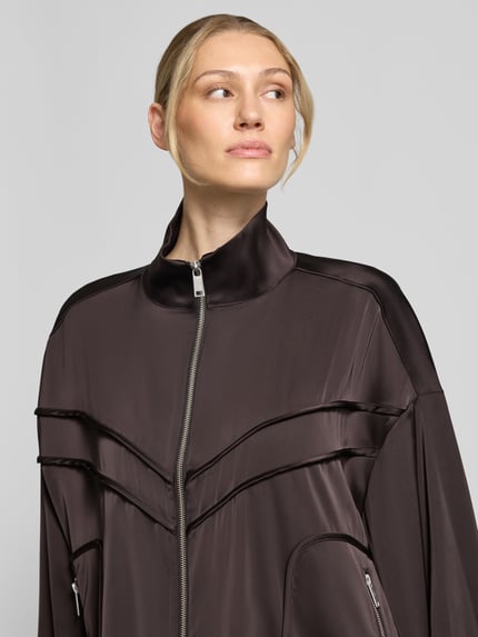 Frauenmodel trägt Relaxed Fit Jacke mit Stehkragen Modell 'Darzi' von Gestuz, dunkler Braunton, glatte Struktur, Nahaufnahme Oberkörper. Elastischer Material-Mix, sichtbarer Reißverschluss, Design mit Ziernähten, Kontext: Rückenlänge 62 cm, Schulterbreite 44 cm, Brustweite 65 cm, Ärmellänge 56 cm.