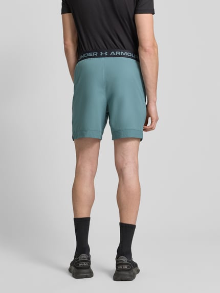 Męski model nosi Szorty z elastycznym pasem i logo od Under Armour, krój regular fit, detale z logo, kolor niebiesko-zielony, widok całej sylwetki z tyłu. Szerokość w pasie rozmiar M: 39 cm, długość wewnętrzna nogawki: 17 cm.