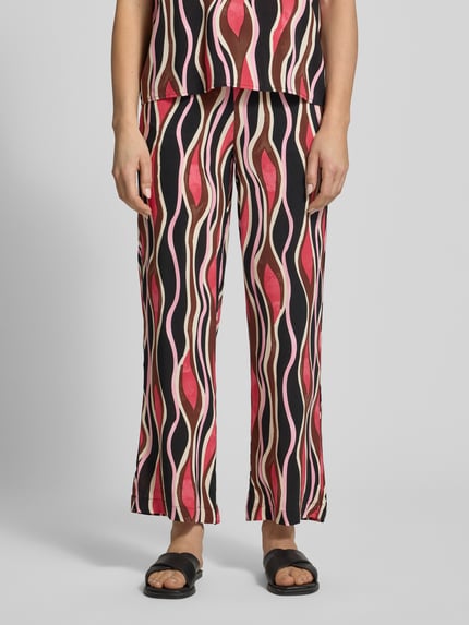 Frauenmodel trägt Straight Fit Stoffhose aus reiner Viskose Modell 'Cammi' von Soyaconcept, grafisches Muster in Rot, Beige und Schwarz, elastischer Bund mit Tunnelzug, Bundweite bei Größe S 35 cm, Innenbeinlänge 74 cm, Ganzkörperansicht von vorne.