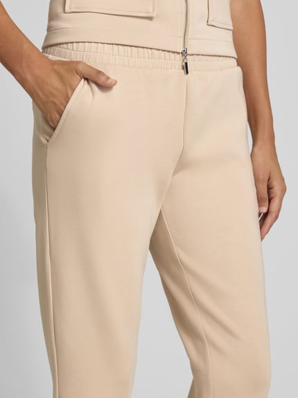 Kobieta nosi Cropped Sweatpants mit elastischem Bund und Eingrifftaschen od Christian Berg Woman, beżowe, miękki materiał, zbliżenie pasa i kieszeni, materiał z domieszką modalu, detal ubrania, zbliżenie okolicy bioder.