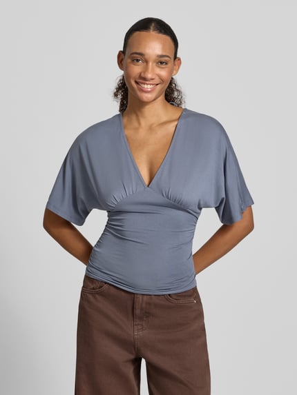 Frauenmodel trägt Bluse aus Viskose-Mix mit V-Ausschnitt von Gina Tricot in Blau, taillierte Passform, Halbarm, Ganzkörperansicht von vorne.