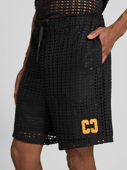 Männermodel trägt Straight Fit Shorts in Loch-Optik von COLUCCI, schwarzer elastischer Material-Mix, gelbes Logo-Stitching, Nahaufnahme Oberschenkel.