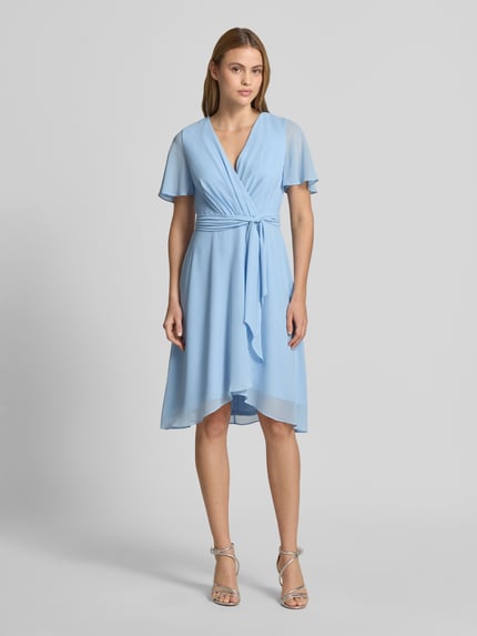 Frauenmodel trägt Knielanges Cocktailkleid in Wickel-Optik von Christian Berg Cocktail aus blauem Chiffon mit Bindegürtel, A-Linie, V-Ausschnitt, Ganzkörperansicht von vorne.