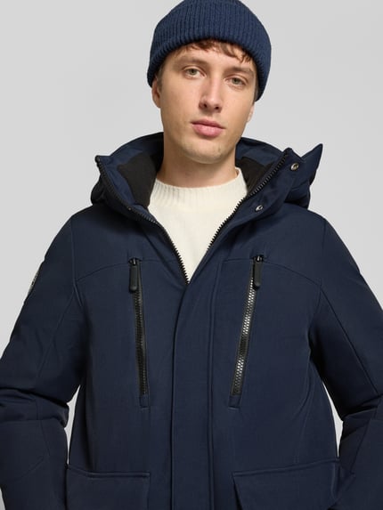 Mężczyzna nosi Parka mit Kapuze und Pattentaschen od Superdry, kolor granatowy, zamek błyskawiczny, krój regular fit, kaptur, kieszenie z patką, zbliżenie górnej części ciała, kontekst: długość tyłu w rozmiarze M: 80 cm, szerokość ramion w rozmiarze M: 48 cm, szerokość klatki piersiowej w rozmiarze M: 61 cm, długość rękawów w rozmiarze M: 73 cm