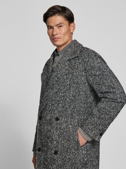 Männermodel trägt Mantel mit Reverskragen von MCNEAL, Fischgrat-Muster, gerade Passform, graue und schwarze Töne, Nahaufnahme Oberkörper.