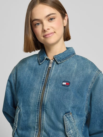 Kobiece model prezentuje Regular Fit Jeansjacke aus reiner Baumwolle von Tommy Jeans w kolorze niebieskim, widoczny label patch, zbliżenie na górną część ciała.