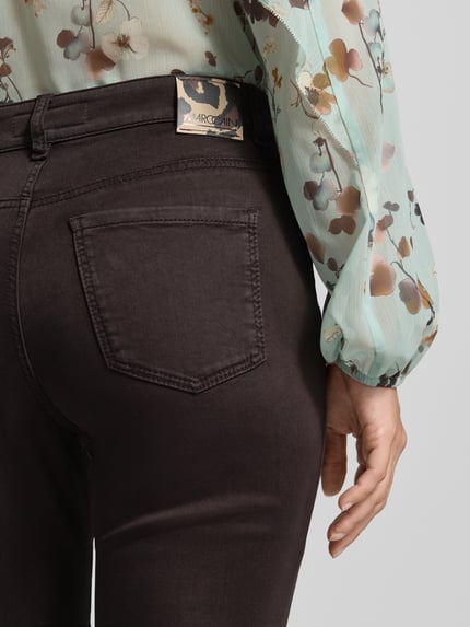 Kobieta nosi Flared Fit Jeans mit Lyocell-Anteil od Marc Cain z elastycznej mieszanki materiałów, 5 kieszeni, zapięcie na guzik i zamek błyskawiczny, szerokość w rozmiarze 36: 37 cm, długość wewnętrzna nogawki w rozmiarze 36: 81 cm, ujęcie: zbliżenie na tył.