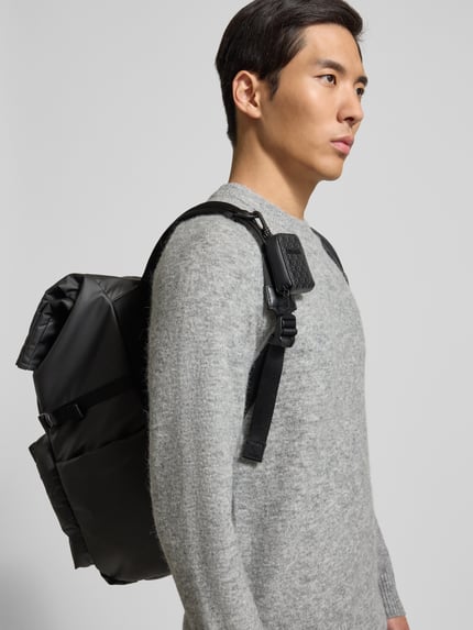 Männermodel trägt Rolltop-Rucksack mit Label-Patch von CK Calvin Klein in Schwarz, Seitenfächer, Klickverschluss, Nahaufnahme Oberkörper