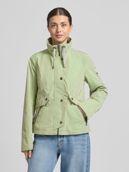 Modelka kobieca nosi Regular Fit Jacke mit Logo-Patch od Gil Bret w jasnozielonym kolorze, gładka tekstura, stehkraag z troczkiem, kieszenie na zamek, zipper z zakładką, długość tyłu 64 cm, szerokość ramion 41 cm, szerokość w rozmiarze 36: 46 cm, długość rękawa 65 cm, widok na cały korpus z przodu.