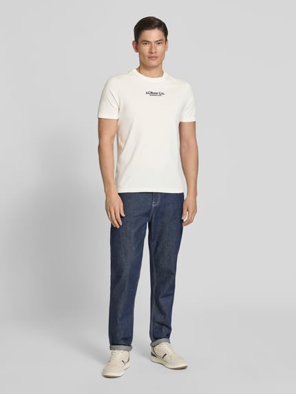 Mężczyzna nosi Regular Fit T-Shirt aus reiner Baumwolle von s.Oliver RED LABEL, biała koszulka z nadrukiem, gładka faktura, krój regular fit, okrągły dekolt, krótkie rękawy, materiał jersey z czystej bawełny; widok całej sylwetki od przodu