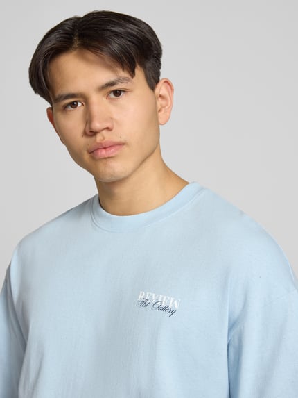 Männermodel trägt T-Shirt mit Label-Detail von REVIEW in Hellblau, Baumwolle, lockerer Schnitt, Nahaufnahme Oberkörper.