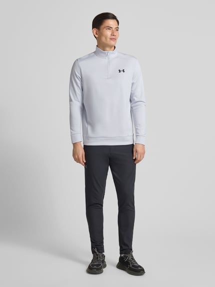 Männermodel draagt Sweatshirt met Labeldetail van Under Armour, los model, opstaande kraag, korte ritssluiting, ribboorden, ruglengte maat M 72 cm, mouwlengte maat M 67 cm. Ganzkörperansicht von vorne.