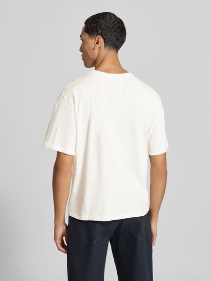 Männermodel trägt Regular Fit T-Shirt aus reiner Viskose von RHUDE in Off-White, glatte Textur, dezenter Label-Print, Ganzkörperansicht Rückansicht. Rückenlänge bei Größe M: 63 cm, Rundhalsausschnitt.