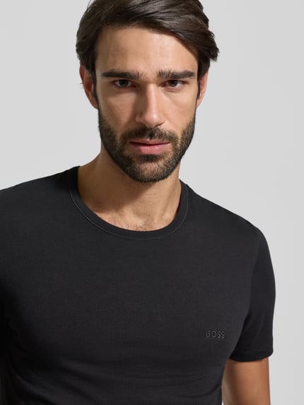 Mężczyzna model nosi T-Shirt mit Label-Stitching im 3er-Pack Modell 'Classic' von BOSS, czysta bawełna, prosty krój, kolor czarny, gładka faktura, naszywka z logo, okrągły ściągacz przy szyi, zbliżenie na górną część ciała.