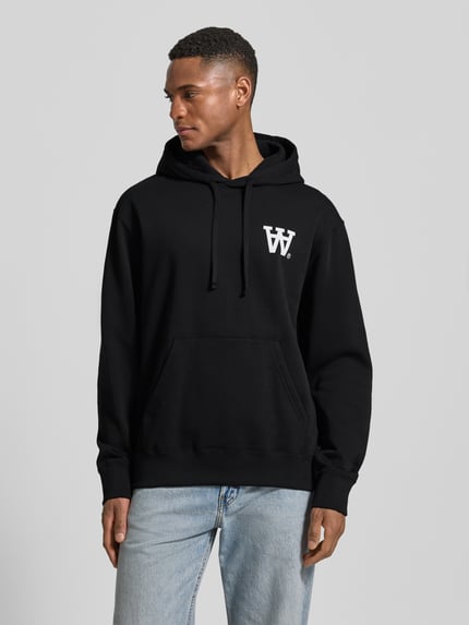 Männermodel trägt Hoodie mit Logo-Print und Känguru-Tasche von Wood Wood, schwarz mit weißem Print, lockere Passform, Ganzkörperansicht von vorne.
