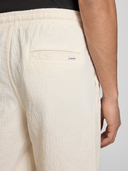 Mężczyzna nosi Shorts mit Strukturmuster Modell 'JAIDEN' od Jack & Jones, kremowy kolor, materiał z bawełną, krój regular fit, tekstura strukturalna, Szerokość w rozmiarze M: 41 cm, długość nogawki wewnętrznej: 21 cm, detal tylnej części, zbliżenie na biodro
