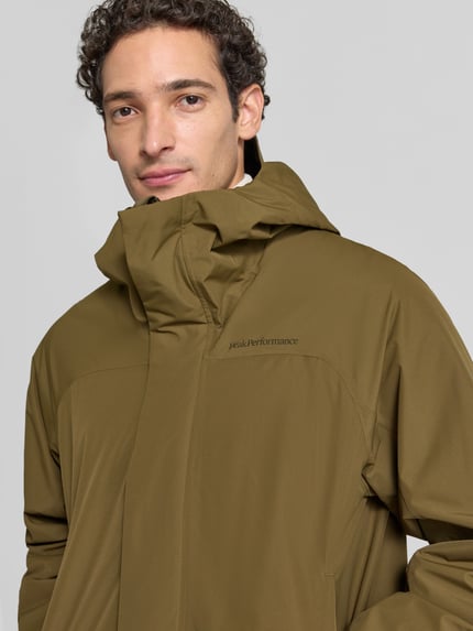Męski model nosi Regular Fit Parka mit Logo Modell 'Treeline HIPE' von Peak Performance, kolor oliwkowy, kaptur, zamek błyskawiczny zakryty, Długość tyłu w rozmiarze M: 100 cm, szerokość ramion w rozmiarze M: 50 cm, szerokość klatki piersiowej w rozmiarze M: 57 cm, długość rękawów w rozmiarze M: 62 cm, z widocznym logo, zbliżenie na górną część ciała