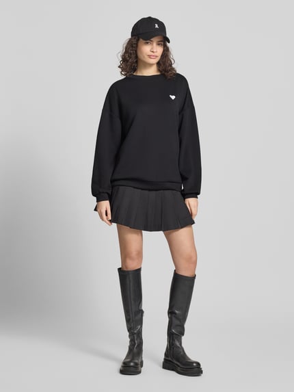 Kobiece model nosi sweter Relaxed Fit Pullover mit Herz-Stickerei Model 'TALIA' od Pieces, efekt: jednolity kolor, krój relaxed fit, wyhaftowane serce, okrągły dekolt, długi rękaw, kolor czarny, gładka faktura, całkowity widok z przodu.