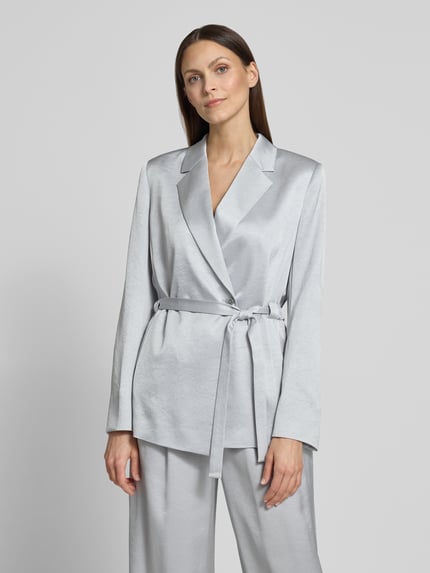 Frauenmodel trägt silbergrauen Satin-Blazer mit Bindegürtel und passender Hose, glatte Oberfläche, Nahaufnahme Oberkörper.