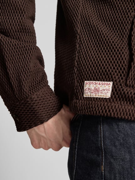 Männermodel trägt Regular Fit Hemdjacke mit Strukturmuster von Scotch & Soda aus Reine Baumwolle in Braun, Detailaufnahme seitlich.