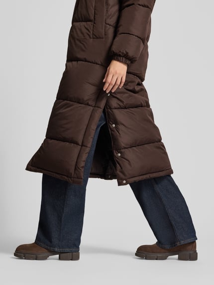 Frauenmodel trägt Puffer Coat mit Kapuze von VERO MODA in Dunkelbraun, Struktur, Regular Fit Longline, sichtbare Teilungsnähte, Detailaufnahme Unterkörper.