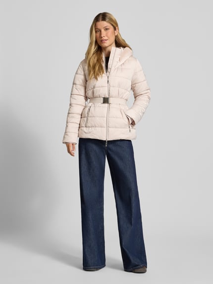 Frauenmodel trägt Steppjacke mit Reißverschlusstaschen und Gürtel von monari in Hellbeige, Kunstfellkapuze, Slim Fit, dezente Steppstruktur, Dunkelblau-Jeans, Ganzkörperansicht von vorne. Rückenlänge 67 cm, Schulterbreite 42 cm, Brustweite 49 cm, Ärmellänge 62 cm.
