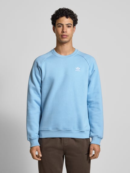 Männermodel trägt Sweatshirt mit Label-Stitching von adidas Originals in Hellblau, Baumwoll-Mix, lockerer Schnitt, gerippte Abschlüsse, Rundhalsausschnitt, Ganzkörperansicht von vorne.