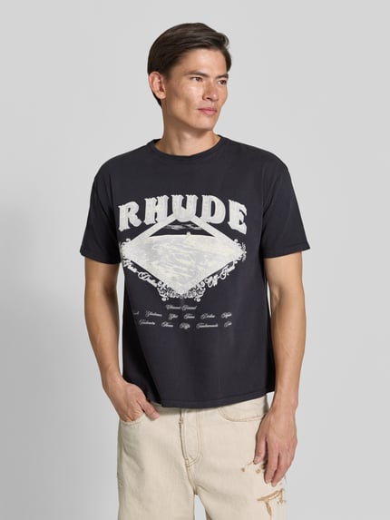 Männermodel trägt Regular Fit T-Shirt aus reiner Baumwolle von RHUDE in Schwarz mit weißem Label-Print, Rundhalsausschnitt, Kurzarm, Ganzkörperansicht von vorne.