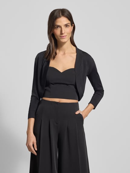 Frauenmodel trägt Bolero mit Schalkragen von Jake*s Cocktail aus schimmernder Viskosemischung mit gerippten Abschlüssen und Dreiviertel-Ärmeln, offene Vorderseite, Rückenlänge 39 cm bei Größe 36, Ganzkörperansicht von vorne.