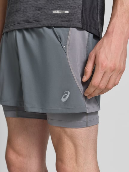 Männermodel trägt Regular Fit Shorts mit integrierten Tights Modell 'Road' von ASICS TIGER in Grau mit Logo-Detail, Nahaufnahme Oberschenkel. Bund mit elastischem Einsatz, glatte Struktur, sportliche Form.