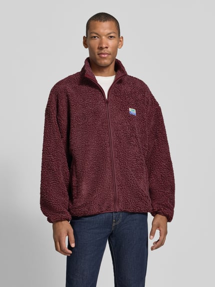 Männermodel trägt Sweatjacke mit Fleece-Struktur Modell 'HOKTOWN' von AMERICAN VINTAGE in Dunkelrot, Reißverschluss, Struktur-Optik, Label-Patch, Ganzkörperansicht von vorne.