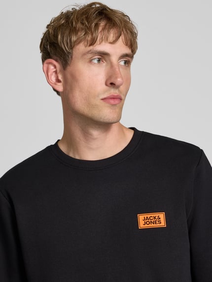 Męskamodel nosi czarną bluzę z mieszanki bawełny o luźnym kroju, nadruk z logo, okrągły dekolt, prążkowane wykończenia, Model 'RUSH' od JACK & JONES, długość tyłu w rozmiarze M: 67 cm, długość rękawów w rozmiarze M: 67 cm, zbliżenie na górną część ciała