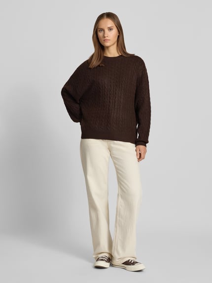 Frauenmodel trägt Oversized Fit Strickpullover mit Zopfmuster von REVIEW in Dunkelbraun, elastischer Material-Mix, helle Hose, Ganzkörperansicht von vorne.