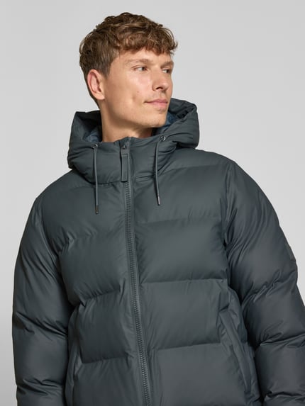 Männermodel trägt Steppjacke mit Zweiwege-Reißverschluss Modell 'Alta' von Rains in Dunkelgrau, glatte Oberfläche, Nahaufnahme Oberkörper. Regular Fit, Kapuze, Rückenlänge 74 cm, Schulterbreite 44 cm, Brustweite 52 cm, Ärmellänge 68 cm.