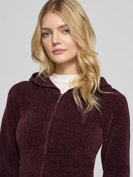 Frauenmodel trägt Sweatjacke mit Kapuze Modell 'MAYA KNIT HOODIE' von Juicy Couture in dunklem, unifarbenem Strick, Logo-Stitching, Nahaufnahme Oberkörper.