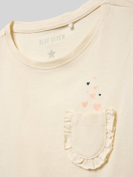 Mädchen trägt Regular Fit T-Shirt mit Rüschenbesatz von Blue Seven aus Reine Baumwolle, Brusttasche, Detailaufnahme Oberkörper.