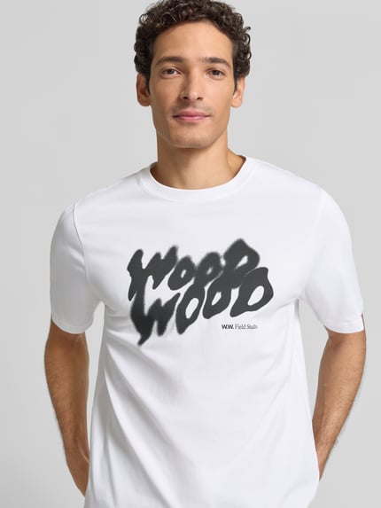 Männermodel trägt T-Shirt mit Label-Print Modell 'Bobby' von Wood Wood, reine Baumwolle, weiß mit schwarzem Print, Nahaufnahme Oberkörper.