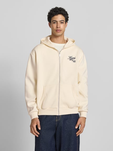 Männermodel trägt Oversized Sweatjacke mit Logo-Stitching von KARL KANI aus Baumwoll-Mix, Kapuze, Kängurutasche, Reißverschluss, cremefarben, Oversized-Fit, elastische Abschlüsse, Rückenlänge 62 cm, Ärmellänge 65 cm, Ganzkörperansicht von vorne.
