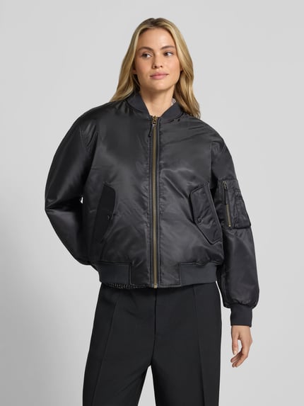 Modelka kobieca nosi Bomberjacke mit Pattentaschen od Anine Bing w kolorze czarnym, luźny krój, zamek na całej długości, stójka, ściągacze, kieszenie z patkami i kieszeń na rękawie; długość pleców w rozm. S: 64 cm, szerokość w rozmiarze S: 60 cm, długość rękawa w rozmiarze S: 60 cm, całopostaciowy widok z przodu.