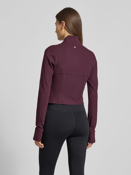 Frauenmodel trägt Slim Fit Jacke mit stójka model 'Storm' od Fabletics, krój krótki, pętelki na kciuki, kolor bordowy, Ganzkörperansicht Rückansicht. Długość tyłu w rozmiarze S: 44 cm, szerokość ramion w rozmiarze S: 36 cm, szerokość klatki piersiowej w rozmiarze S: 42 cm, długość rękawów w rozmiarze S: 74 cm.