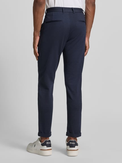 Męski model nosi Slim Fit Hose mit Viskose-Anteil in verkürzter Passform von Lindbergh, ciemnogranatowy kolor, elastyczna mieszanka wiskozy, wpuszczane kieszenie, guzik i zamek błyskawiczny, szerokość w rozmiarze M: 42 cm, długość wewnętrzna nogawki: 72 cm, spodnie o kroju slim fit, skrócony fason, widok całej sylwetki z tyłu.