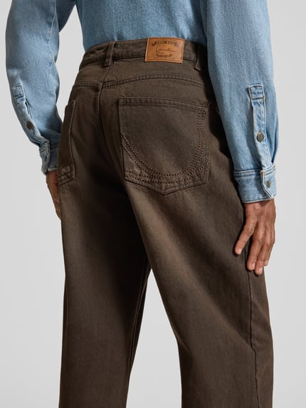 Männermodel trägt Relaxed Fit Jeans mit Knopf- und Reißverschluss von KARL KANI in Braun, 5-Pocket-Design, Label-Patch, Ganzkörperansicht Rückansicht.