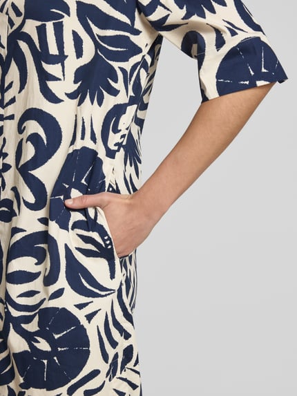 Kobieta nosi Knielanges Hängerkleid mit Tunikakragen od Oui, mieszanka bawełny z dodatkiem streczu, prosty krój, długość do kolan, kołnierz z lamówką, rękaw o dł. 3/4, długość tyłu w rozmiarze 36: 101 cm, szerokość w rozmiarze 36: 51 cm, długość rękawa: 35 cm, wzór granatowo-kremowy, tekstura miękka, detal kieszeni, zbliżenie na bok sukienki, detalaufnahme.