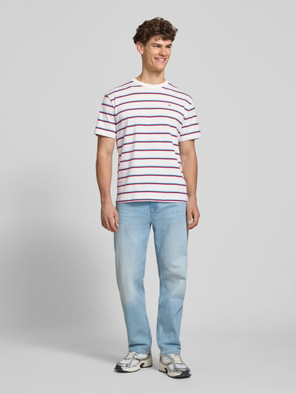 Männermodel trägt Regular Fit T-shirt uit pure katoen van Tommy Jeans met all-over motief, gestreept patroon, logo-stitching, jersey materiaal, ronde hals, korte mouw, heren look. Ganzkörperansicht von vorne.