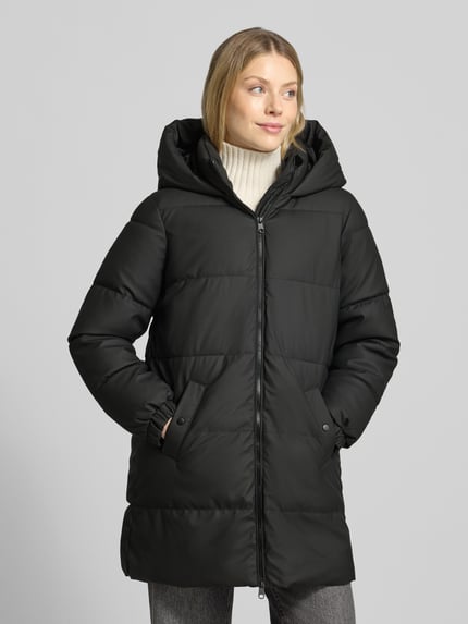 Frauenmodel trägt schwarzen Puffer Coat mit Kapuze von VERO MODA, Struktur mit Teilungsnähten und seitlichen Taschen, Ganzkörperansicht von vorne.